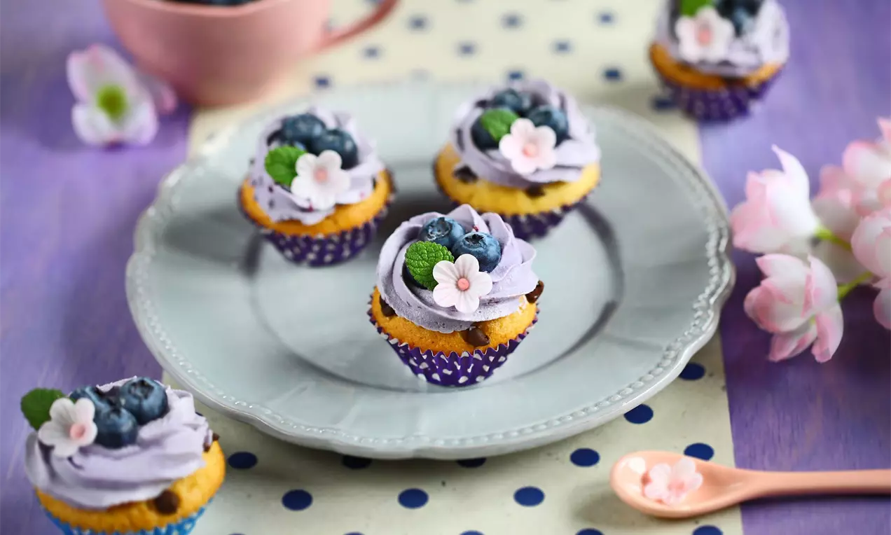 Vanilkové mini muffiny s čokoládovými kousky zdobené fialovým krémem, jedlými kvítky a čerstvými borůvkami na bílém talíři