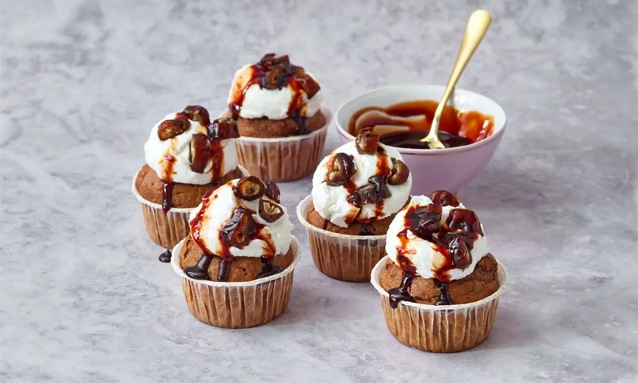 Ovesné muffiny tmavě hnědé barvy s vrstvou šlehačky, zdobené karamelovou polevou a kousky datlí, uspořádané na světlém povrchu