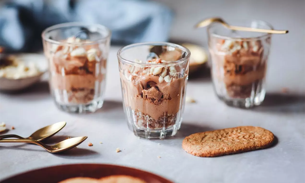 Krémový dezert s čokoládovým mousse ve skleničkách, zdobený nasekanými arašídy, se zlatými lžičkami a sušenkami na podkladu.