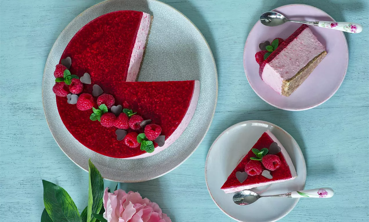 Malinový cheesecake s lesklou červenou polevou, zdobený malinami, lístky máty a čokoládovými srdíčky, servírovaný na světle modrém pozadí
