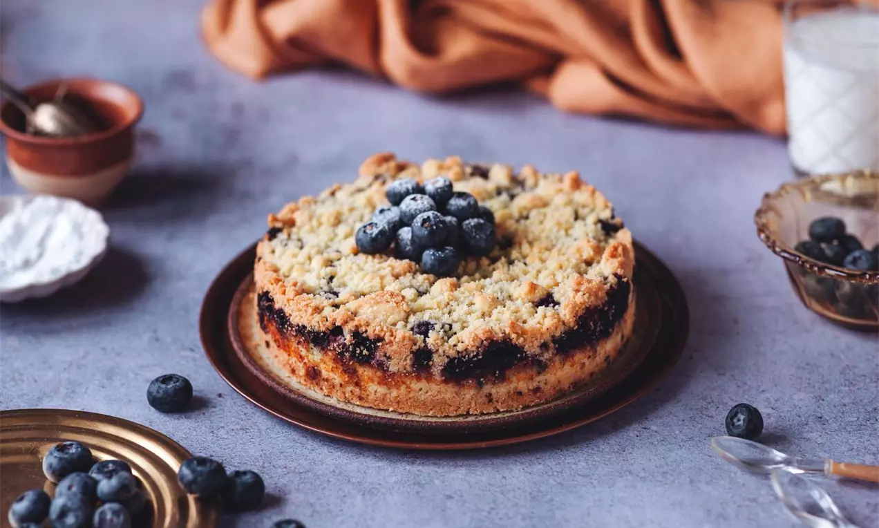 borůvkový koláč s drobenkou zdobený borůvkami na modré pracovní ploše