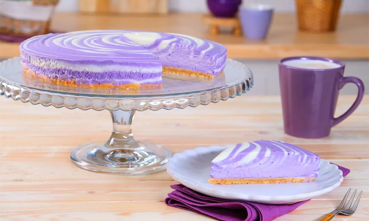 Ube cheesecake s hladkou fialovou náplní s jemnými spirálami bílé barvy, na zlatohnědém sušenkovém základu, servírovaný na skleněném podnosu