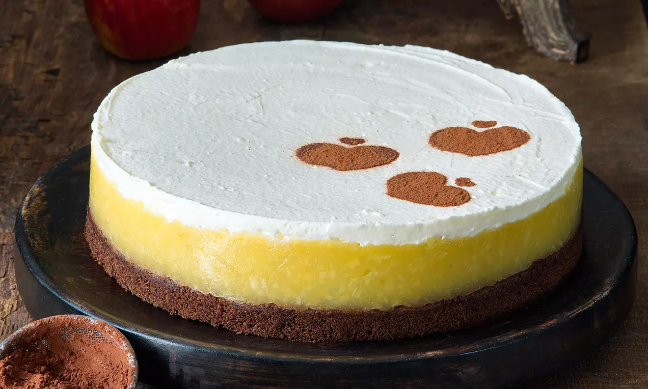 Kulatý jablečný dort na tmavém sušenkovém základu, vrstvený žlutou jablečnou náplní a bílým mascarpone krémem, ozdobený obrázky jablek z kakaa