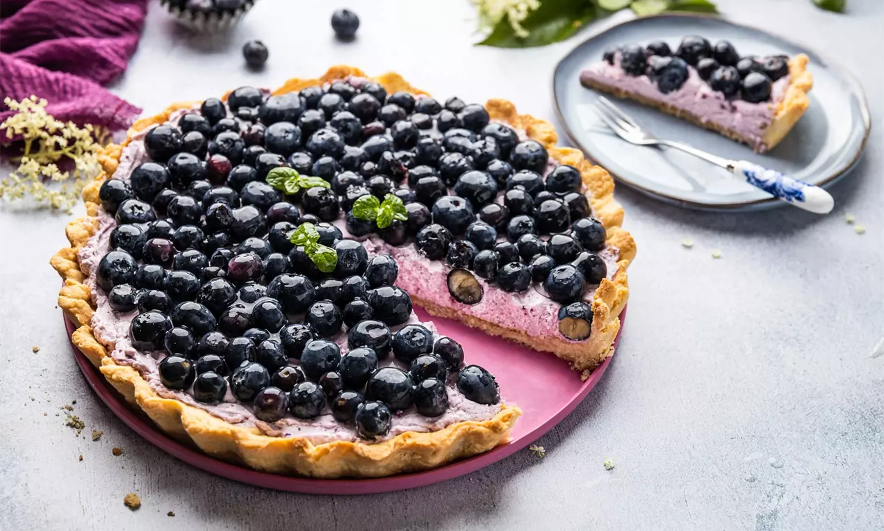 Naříznutý koláč z křehkého těsta s fialovou borůvkovou náplní zdobený čerstvými borůvkami na růžovém servírovacím talíři a bílé pracovní desce