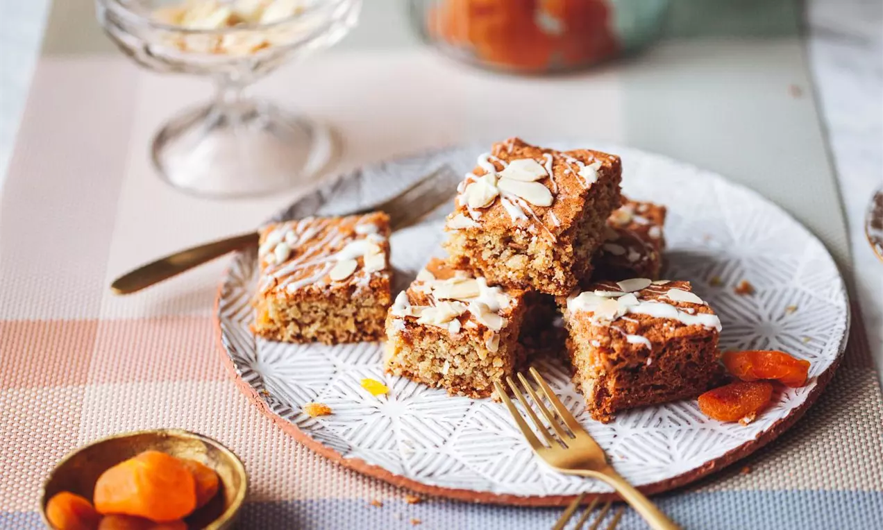 Blondie řezy servírované na šedém talíři se zlatými příbory zdobené plátky mandlí, bílou čokoládou a sušenými meruňkami