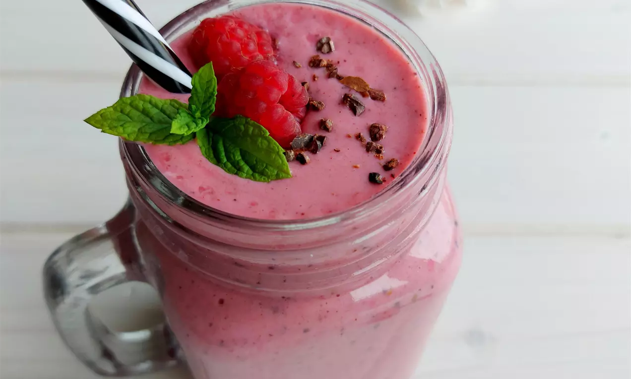 Světlé malinové smoothie v průhledné sklenici s černobílým brčkem, zdobené malinami, mátovými lístky a kakaovými boby