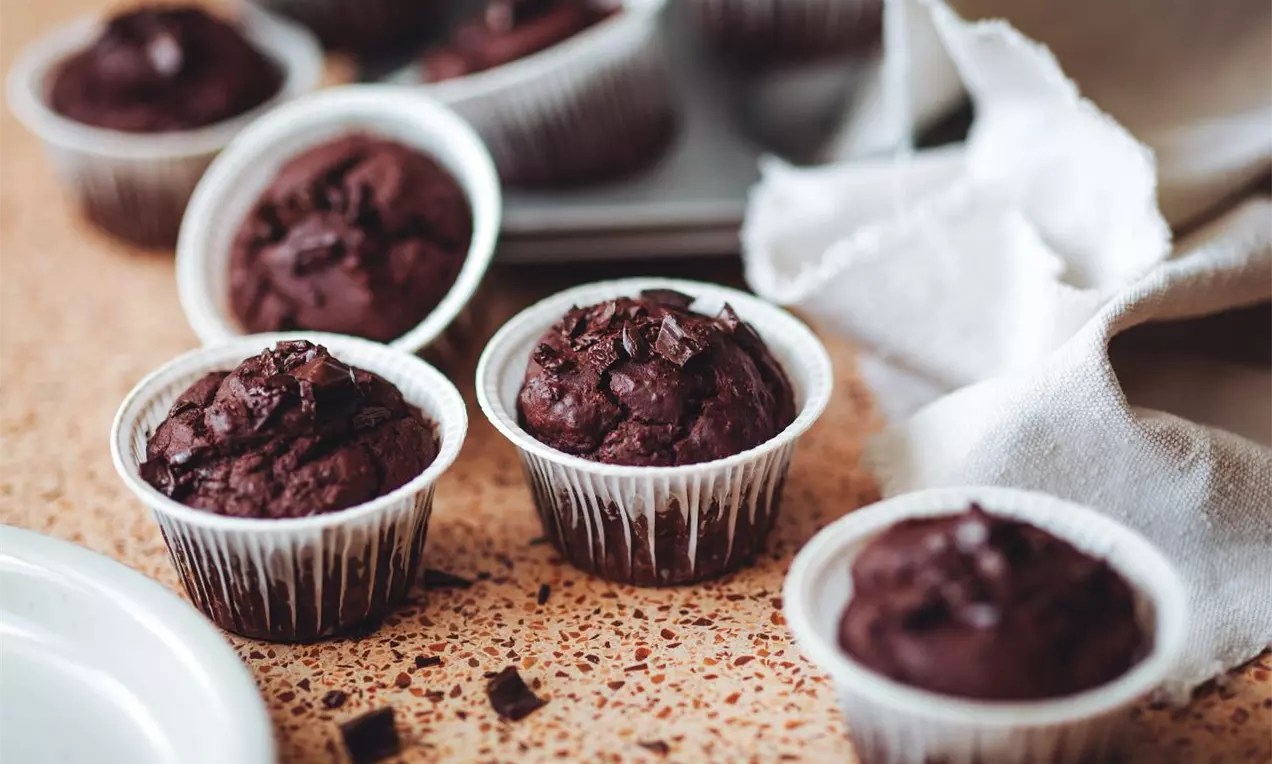 Čokoládově hnědé muffiny s kousky čokolády v bílých papírových košíčcích na světlém zrnitě vzorovaném povrchu vedle bílé látky