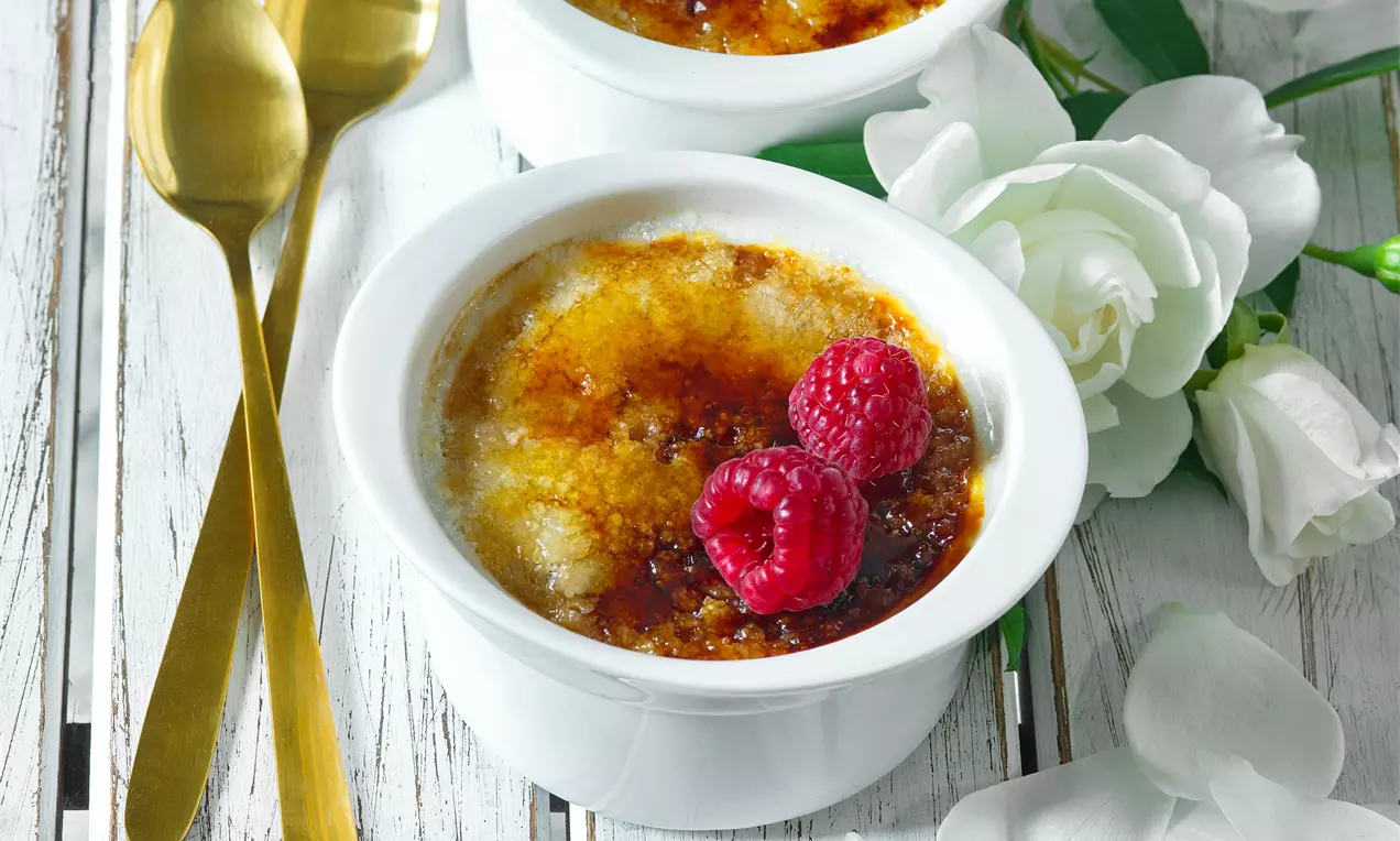 Crème brûlée s karamelově zlatavou krustou nalomenou uprostřed, ozdobené sytě červenými malinami v bílém keramickém ramekinu na dřevěném stole