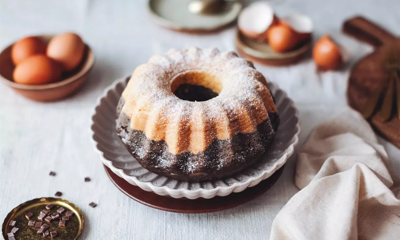 Bábovka ze světlého a tmavého těsta pocukrovaná moučkovým cukrem a pracovní ploše s vajíčky a válečkem v pozadí