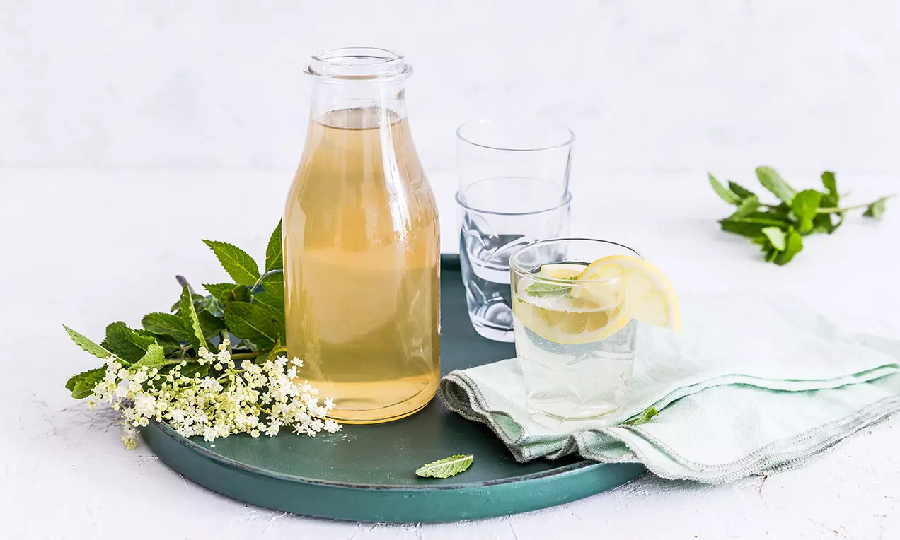 Světle zlatavý sirup v průhledné lahvi s jemným leskem, doplněný sklenicí vody s plátkem citronu a lístkem máty na zeleném podnose