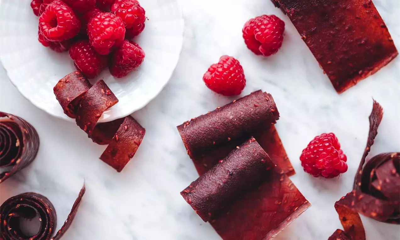 červené plátky z lisovaného ovoce srolované do rolky na bílé mramorové pŕacovní ploše s čerstvými malinami v okolí