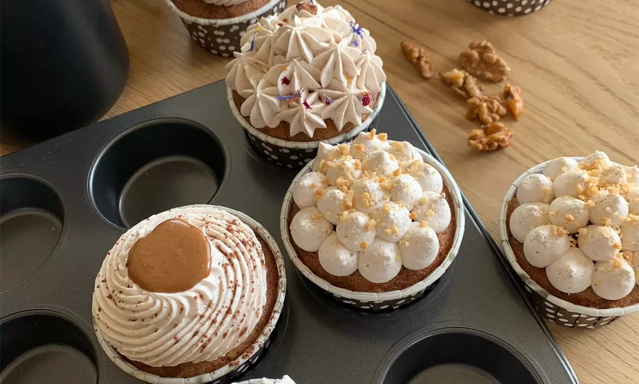 Jednotlivé hnědé muffiny v černobílých papírových košíčcích, zdobené krémovými kopci, ořechy, sušenkou a jedlými květy na plechu