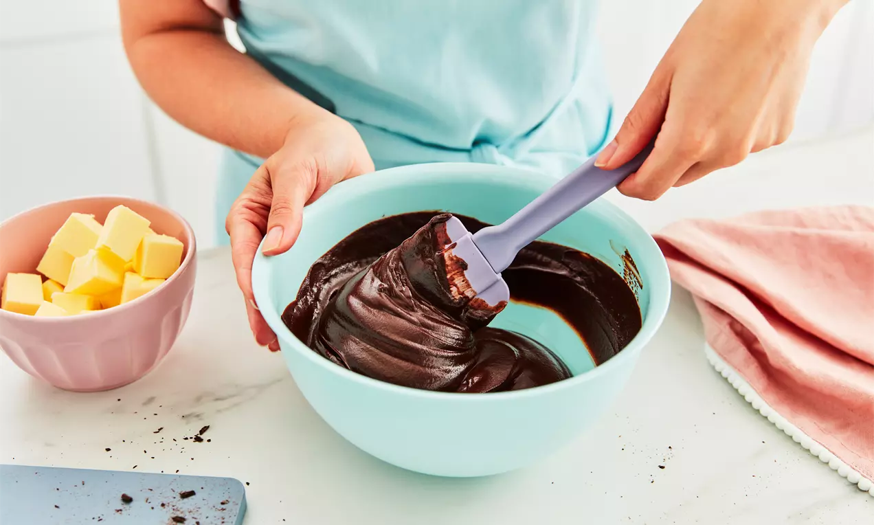 Ruce míchají hladkou tmavou čokoládovou ganache pomocí silikonové stěrky v míse. Vedle jsou připravené kostky másla, které dodávají ganache jemnou konzistenci a lesklý vzhled.