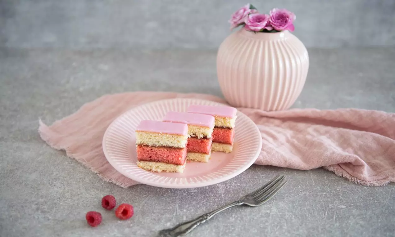 Malinový punčák tvořený vrstvami růžové a světlé piškotové hmoty, přelitý růžovou polevou na jemném růžovém talíři s malinami a vidličkou