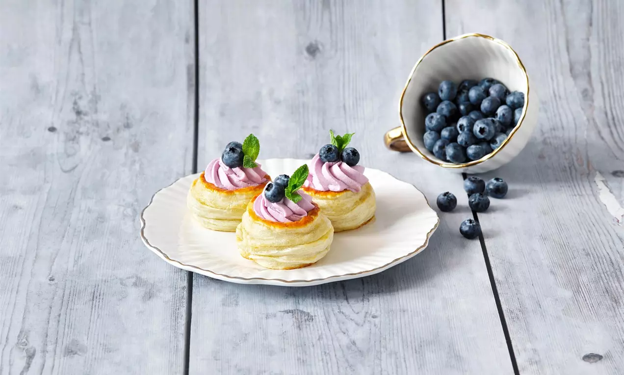 Nadýchané zlatavé pastry s jemným fialovým krémem, borůvkami a lístky máty na světlém talíři vedle šálku plného čerstvých borůvek
