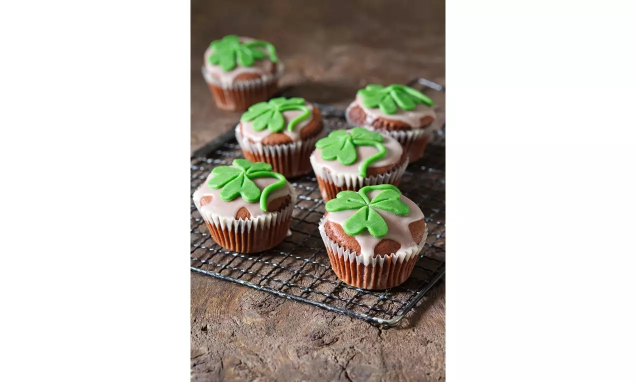 Muffiny s hnědým těstem, světlou marcipánovou polevou, zdobené zelenými čtyřlístky, na drátěné mřížce položené na rustikálním stole