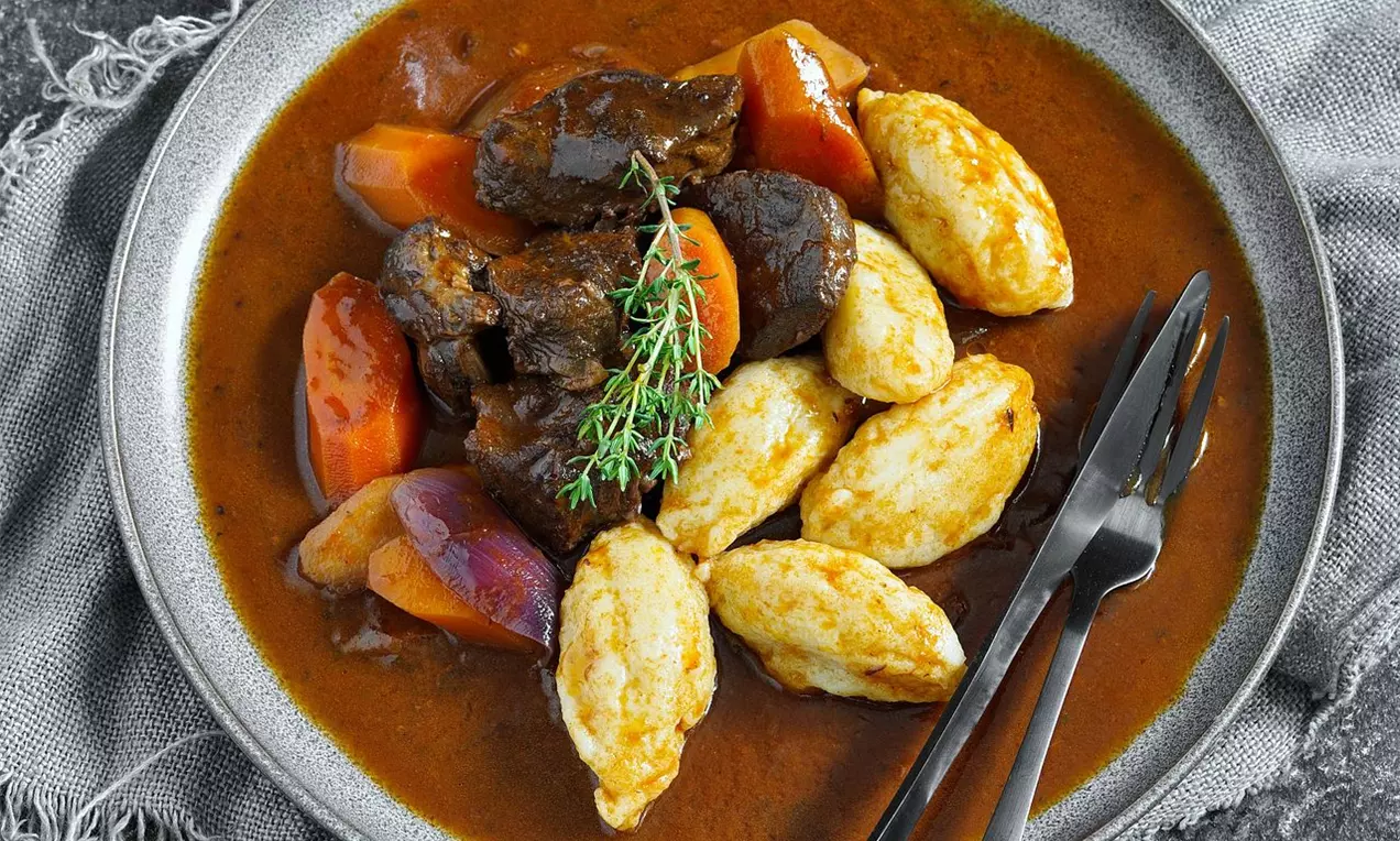 Na šedém talíři hovězí kostky v hnědé omáčce, světlé noky ze zlatožlutého těsta, měkké plátky mrkve, větvička tymiánu a příbor