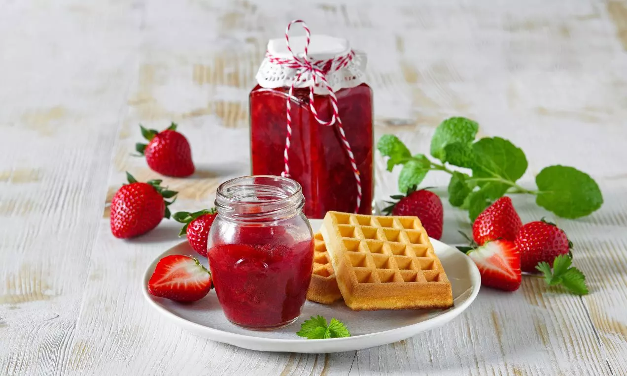 Sklenice s jasně červenou jahodovou marmeládou vedle čerstvých jahod, mátových lístků a zlatavých vaflí na světlém dřevěném podkladu