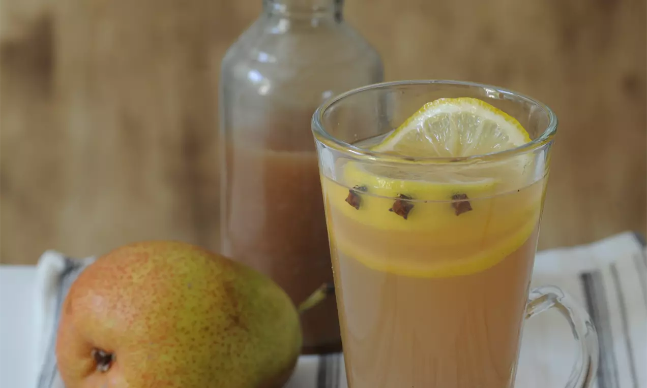 Horký hruškový nápoj světle hnědé barvy ve skleněném hrnku, ozdobený plátkem citronu a hřebíčkem, vedle lahve, skořice a čerstvé hrušky