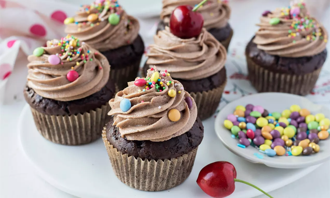 Tmavě hnědé čokoládové cupcakes s krémovou čokoládovou špičkou, zdobené barevným sypáním, třešní a podávané na bílém talíři