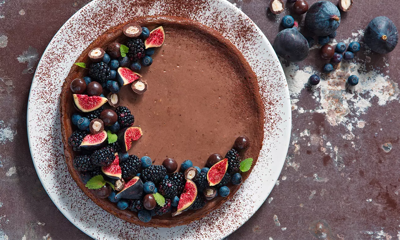 Kakaový cheesecake s hladkým čokoládovým povrchem na bílé talíři, zdobený fíky, ostružinami, borůvkami, lístky máty a kuličkami čokolády