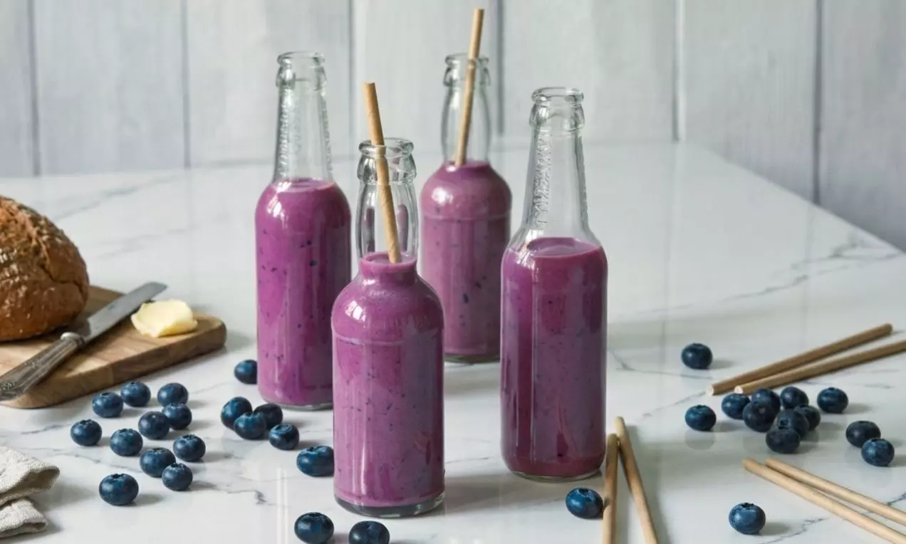 Štíhlé skleněné lahve s hustým fialovým smoothie, doplněné bambusovými brčky, obklopené čerstvými borůvkami na mramorovém stole