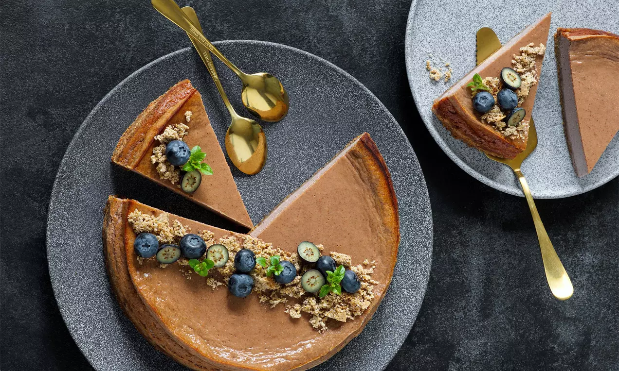 Makový perníkový cheesecake s hladkým lesklým povrchem zdobený borůvkami, drobenkou a lístky máty na šedých talířích s kovovými lžícemi