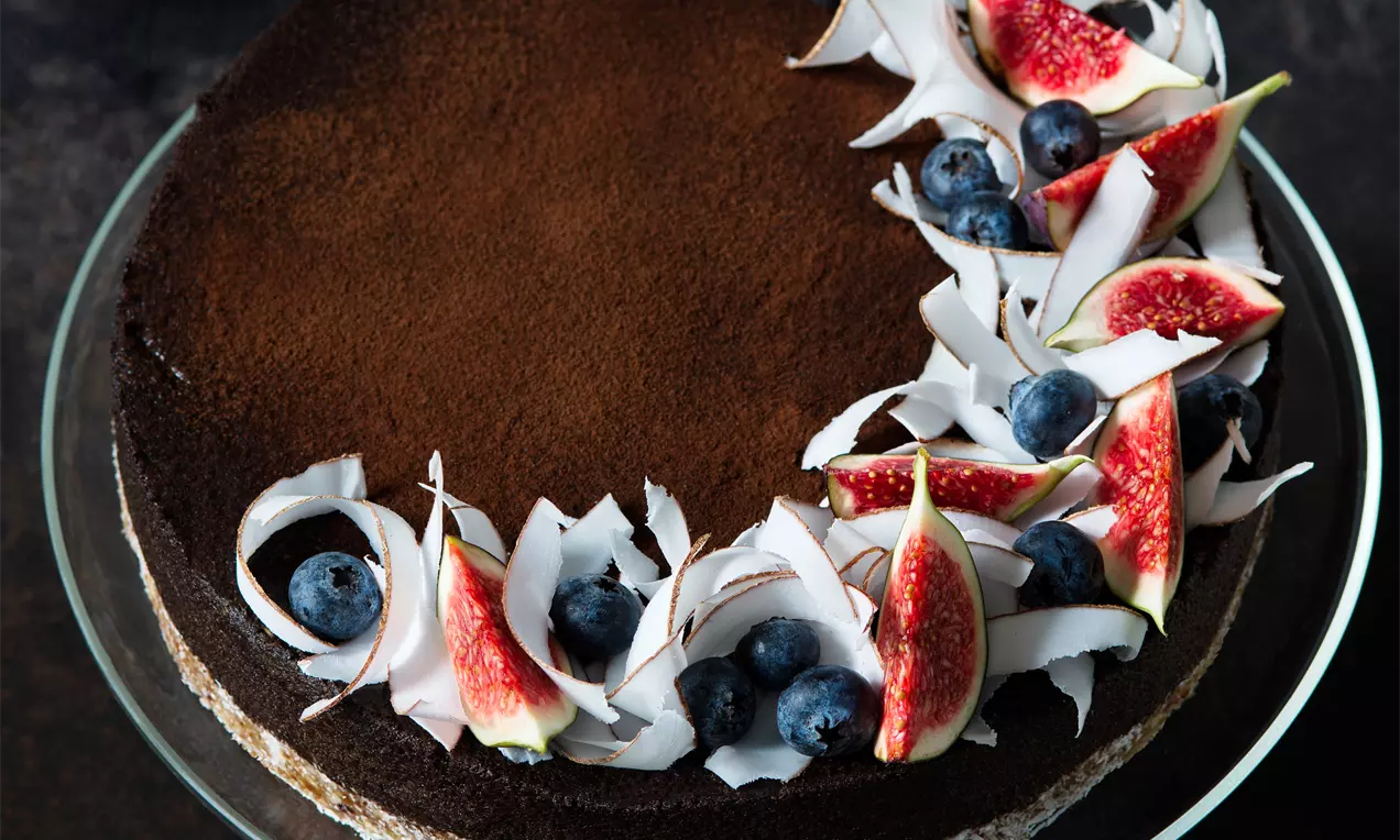 Čokoládově hnědý karobový cheesecake sypaný kakaovým práškem, ozdobený fíky, borůvkami a tenkými kokosovými plátky na skleněném talíři