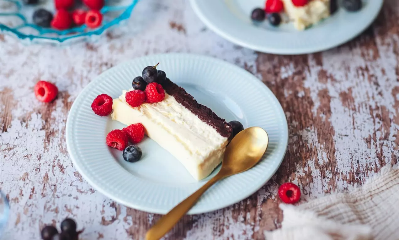 řez cheesecakeu s tmavým korpusem a bílým krémem na modrém dezertním talířku s okolím zdobeným čerstvým lesním ovocem