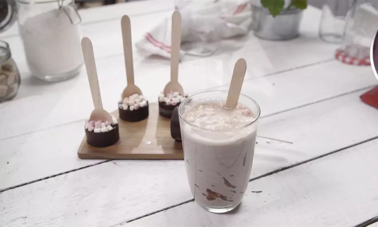 Kostky tmavé čokolády s posypem drobných bílých a růžových marshmallows na dřevěné lžičce vedle sklenice s krémovým nápojem na bílém stole