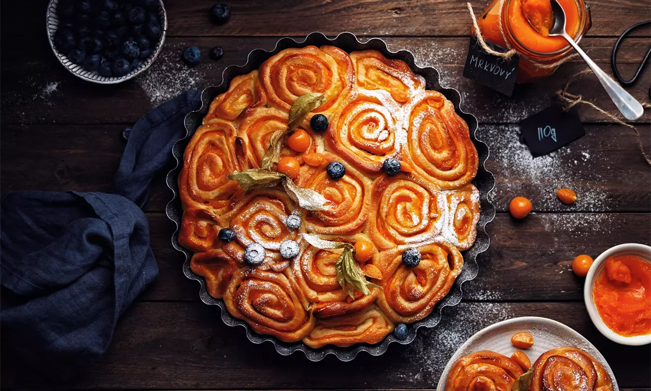 Vosí hnízda zlatohnědá a lesklá, tvořící spirálové vzory v kulaté formě, zdobená mrkvovým džemem, physalis a borůvkami na dřevěném stole