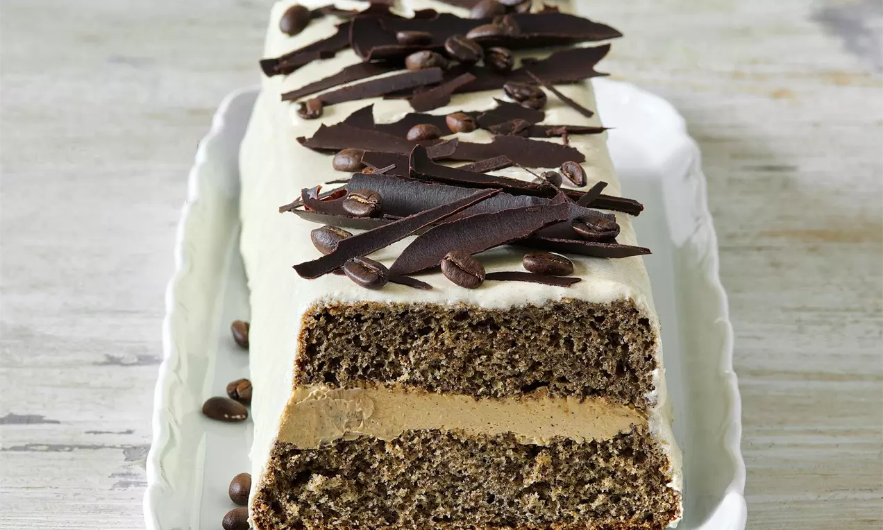 Biskupský chlebíček plněný kávovým krémem politý bílkovou polevou a zdobený čokoládovými hoblinami a kávovými zrnky, na bílém servírovacím talíři a bílém dřevěném stole