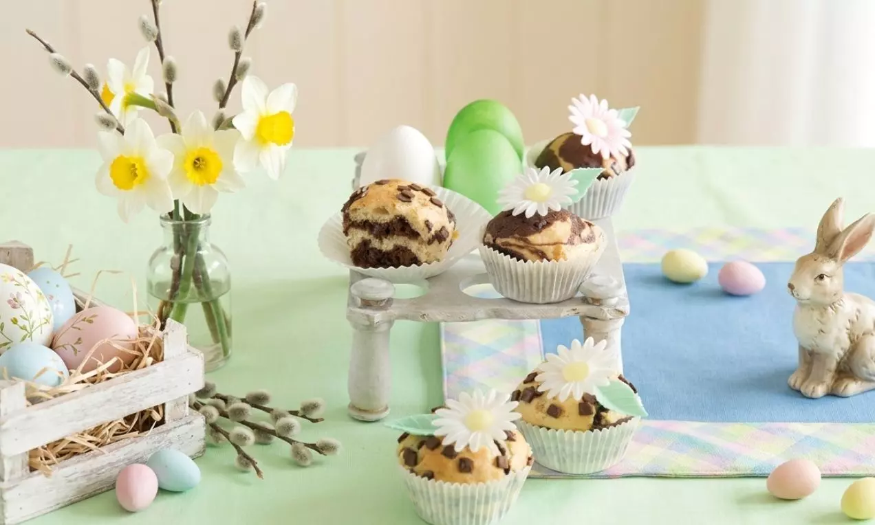 Dekorativní muffiny mramorovaného vzhledu v papírových košíčcích, zdobené bílými květy se zelenými lístky na pastelově laděném stole