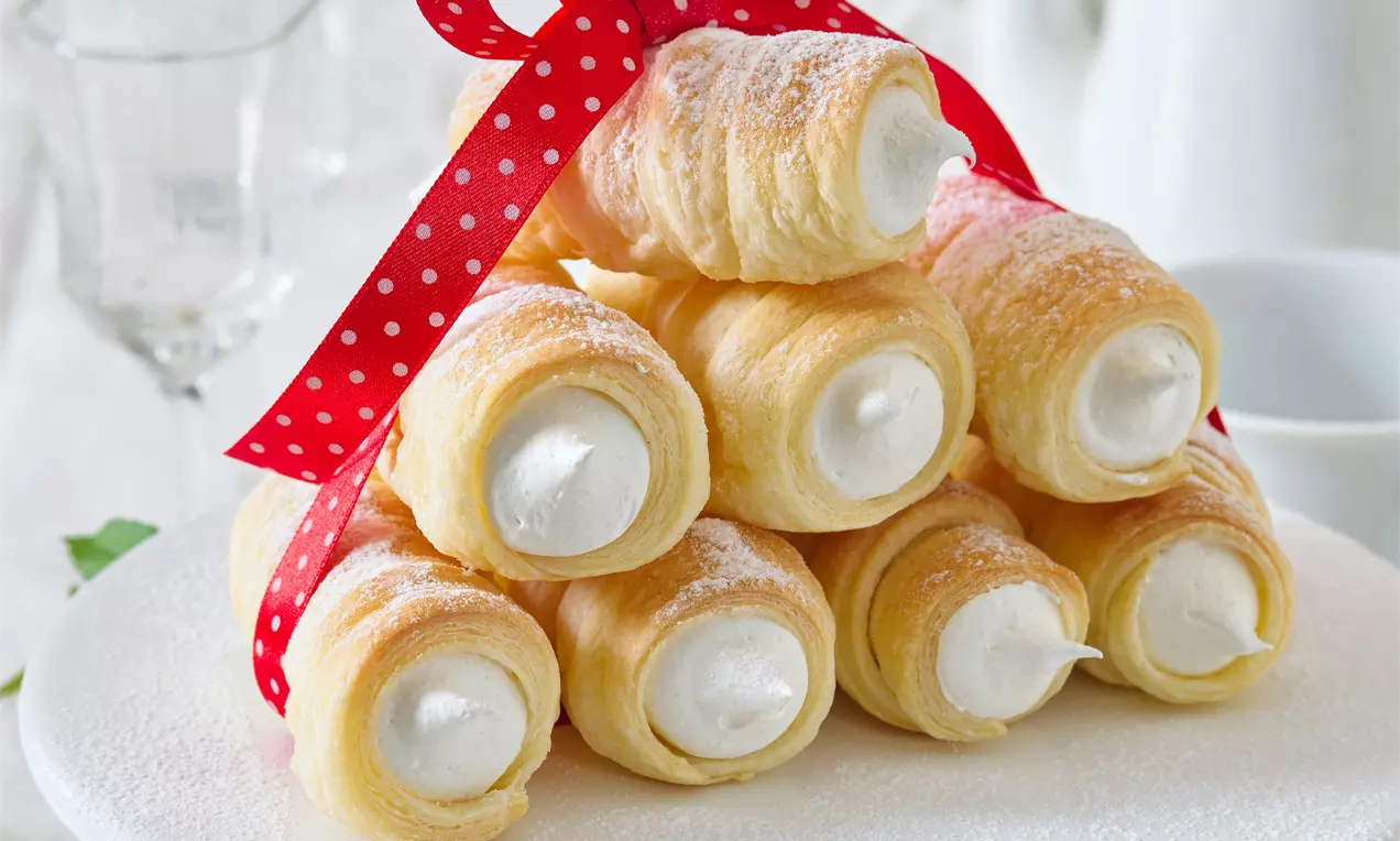 Křehké zlatavé kremrole plněné bílým nadýchaným citronovým sněhem, posypané moučkovým cukrem, na talíři dekorované červenou mašlí