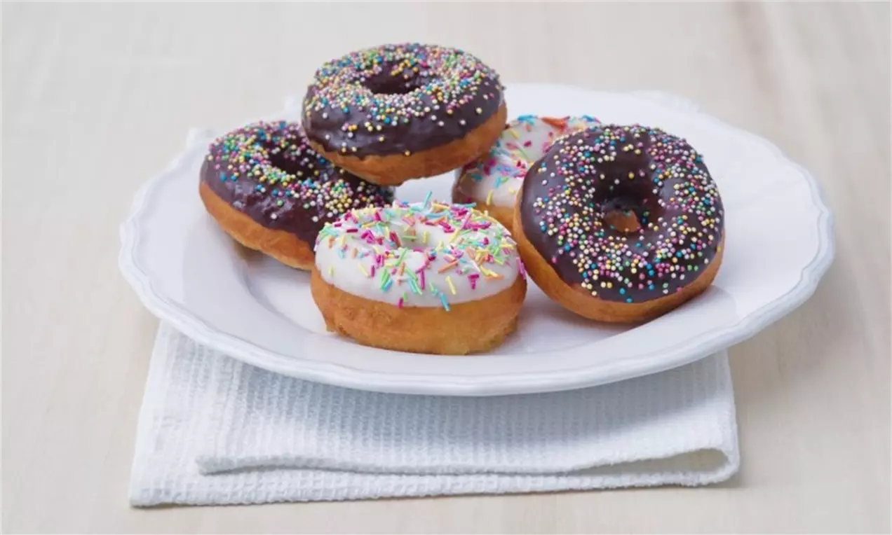 Donuty s hnědou čokoládovou i bílou polevou, posypané barevným cukrovým zdobením, na bílém talíři na látkovém ubrousku