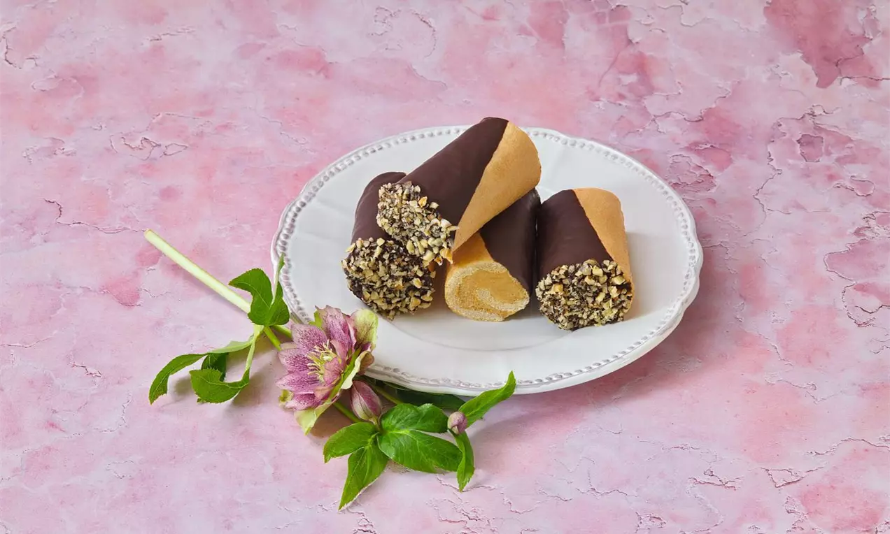 Štafetky zdobené tmavou čokoládou a nasekanými oříšky, na bílém talíři s fialovým květem a zelenými lístky na růžovém pozadí