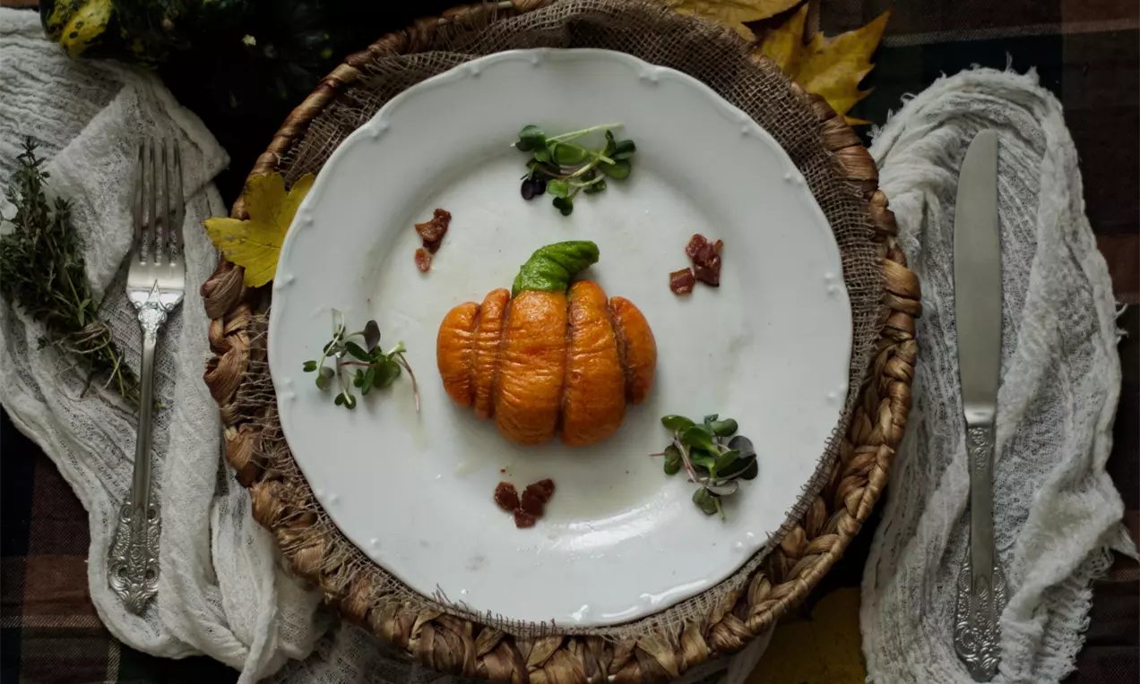 Dýňová buchta oranžová, tvarovaná jako dýně, s jemně lesklým povrchem, obklopená kousky slaniny a microgreens na bílém talíři s rustikálním podkladem