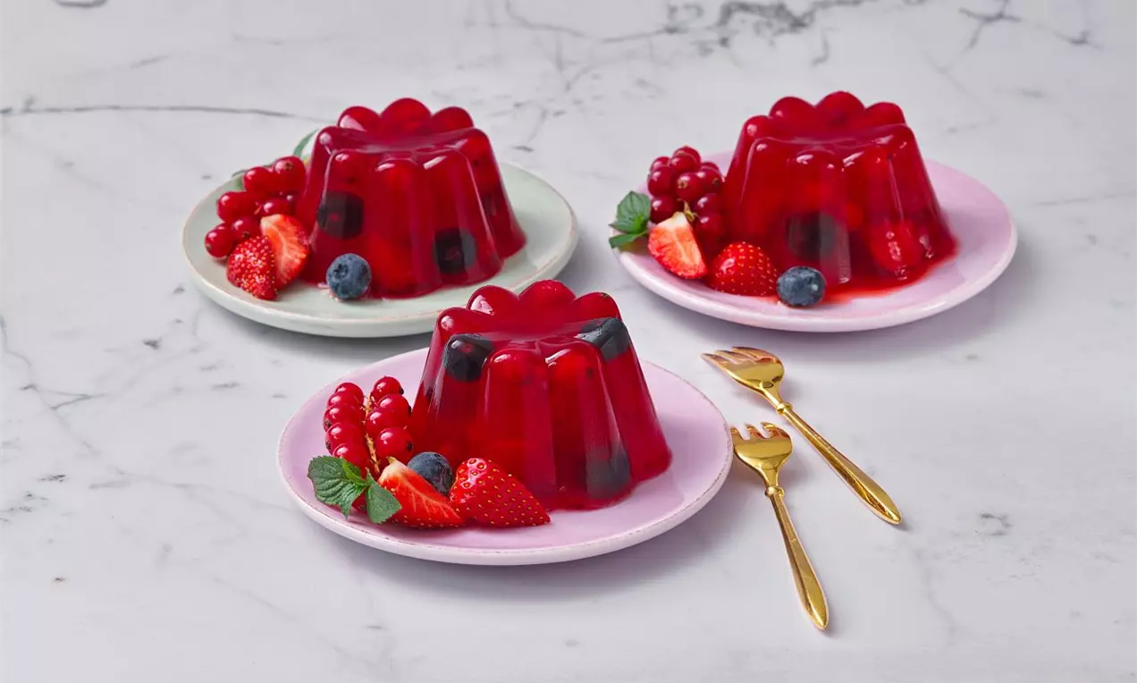 Trojice červených želé báboviček s lesklým povrchem na pastelových talířích, doplněná čerstvými jahodami, rybízem a borůvkami