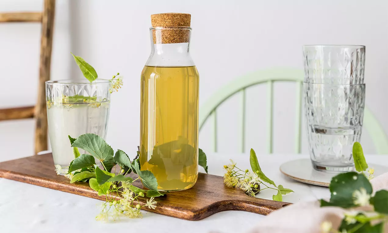 Lipový sirup zlatavé barvy v láhvi s korkovým uzávěrem na dřevěném prkénku obklopeném čerstvými lipovými květy a zelenými lístky