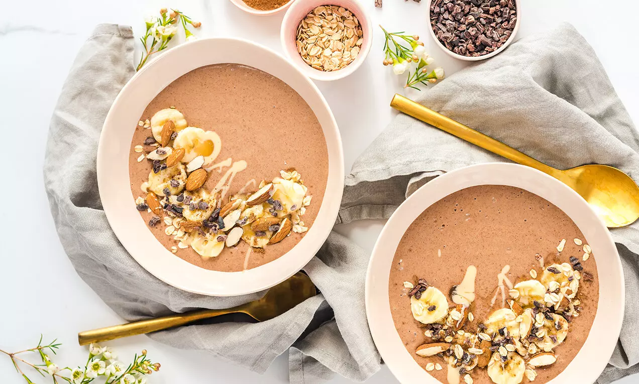 Banánová smoothie miska s krémově hnědým povrchem, zdobená plátky banánu, oříšky, granolou a kapkami krému, naservírovaná na světlém podkladu