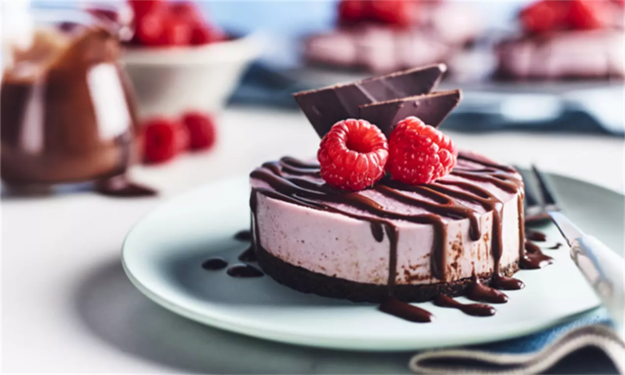 Veganský malinový cheesecake světle růžové barvy na tmavé sušenkové základně, zdobený čokoládovými plátky, malinami a čokoládovým přelivem
