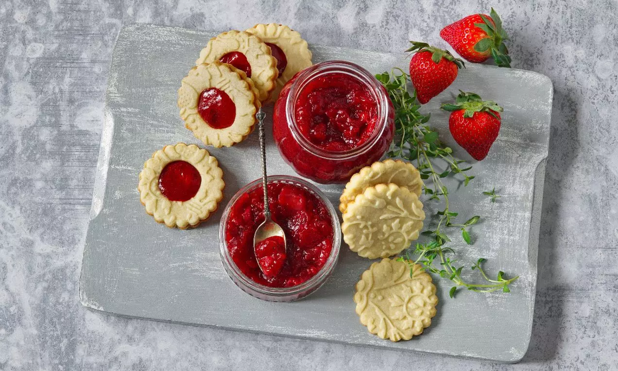 Jahodovo-tymiánová marmeláda zářivě červené barvy v prosklené sklenici s kovovou lžičkou, obklopená sušenkami a čerstvými jahodami na šedém podnosu