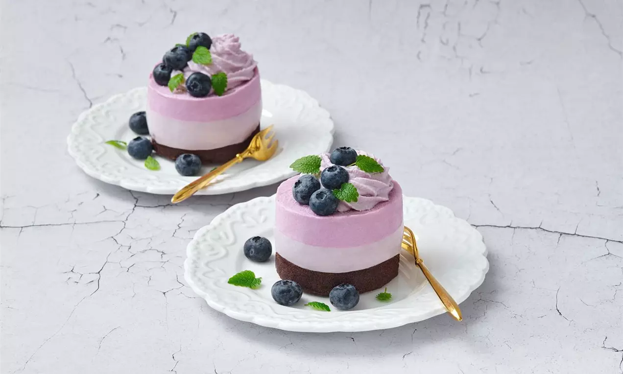 Světlé fialové borůvkové dortíky na tmavém sušenkovém základu, ozdobené čerstvými borůvkami, lístky máty a servírované na bílých talířích