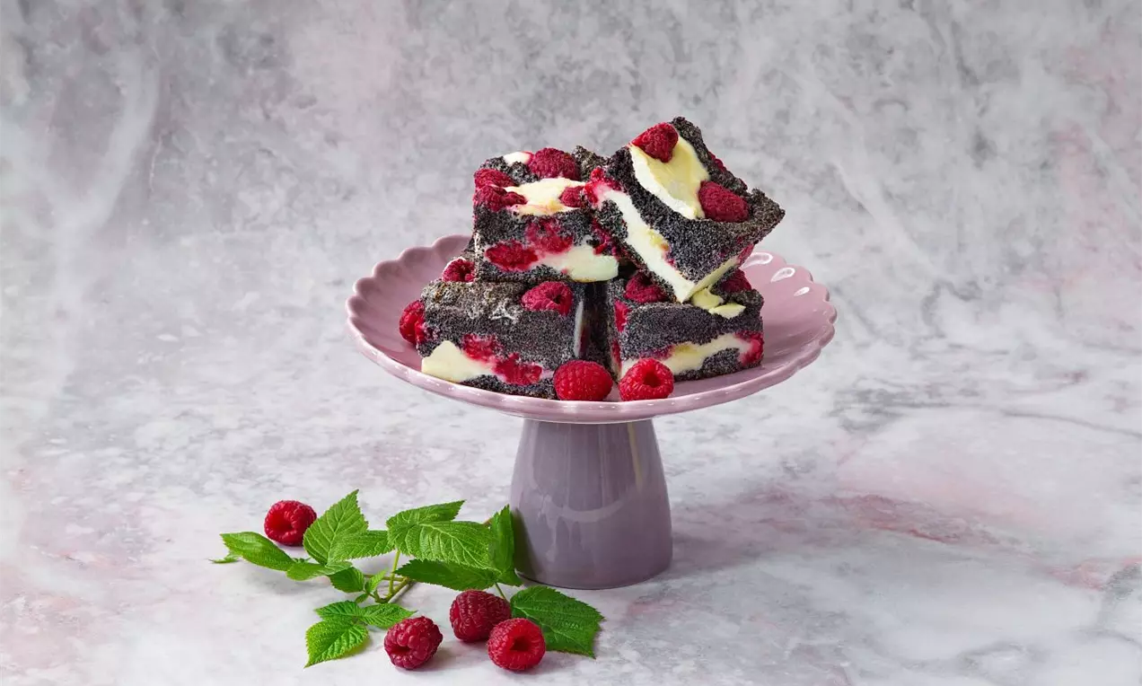 Čtvercové kousky tmavého makového koláče s vrstvičkami světlého krému, ozdobené malinami na růžovém podnosu, vedle čerstvé větvičky malinovníku