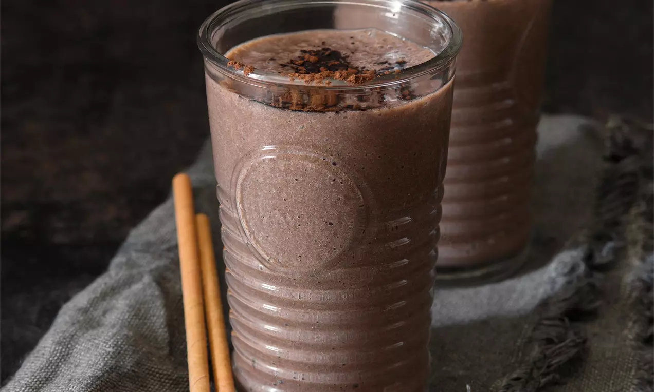 Hnědé smoothie jemné textury ve vysoké sklenici s reliéfem, posypané tmavým práškem, vedle plátěného ubrousku a dvou tyčinek