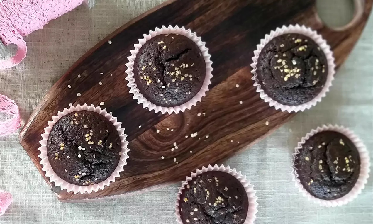 Tmavě hnědé muffiny s jemnou popraskanou texturou, posypané světlými semínky, v růžových papírových košíčcích na dřevěném podnosu