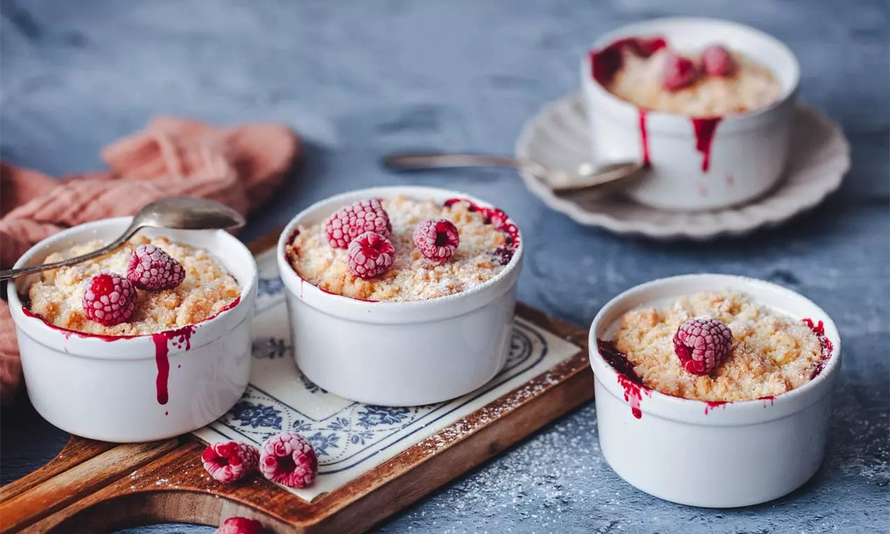 4 misky se zapečeným dezertem s drobenkou na vrchu na dřevěném prkénku a modré pracovní ploše zdobené malinami