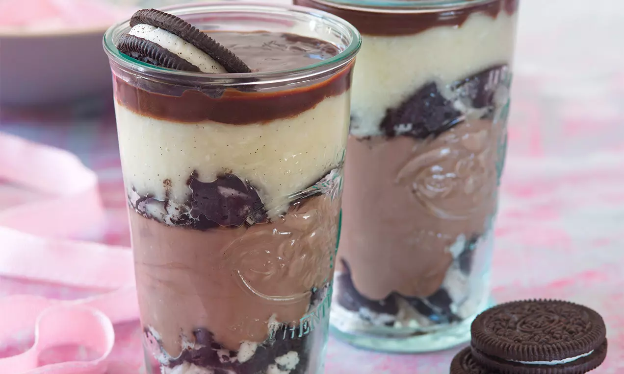 Vysoké skleněné poháry vrstvené tmavohnědými cookies, krémově hnědými a vanilkovými pudinky, přelité čokoládou a zdobené sušenkou na vrchu