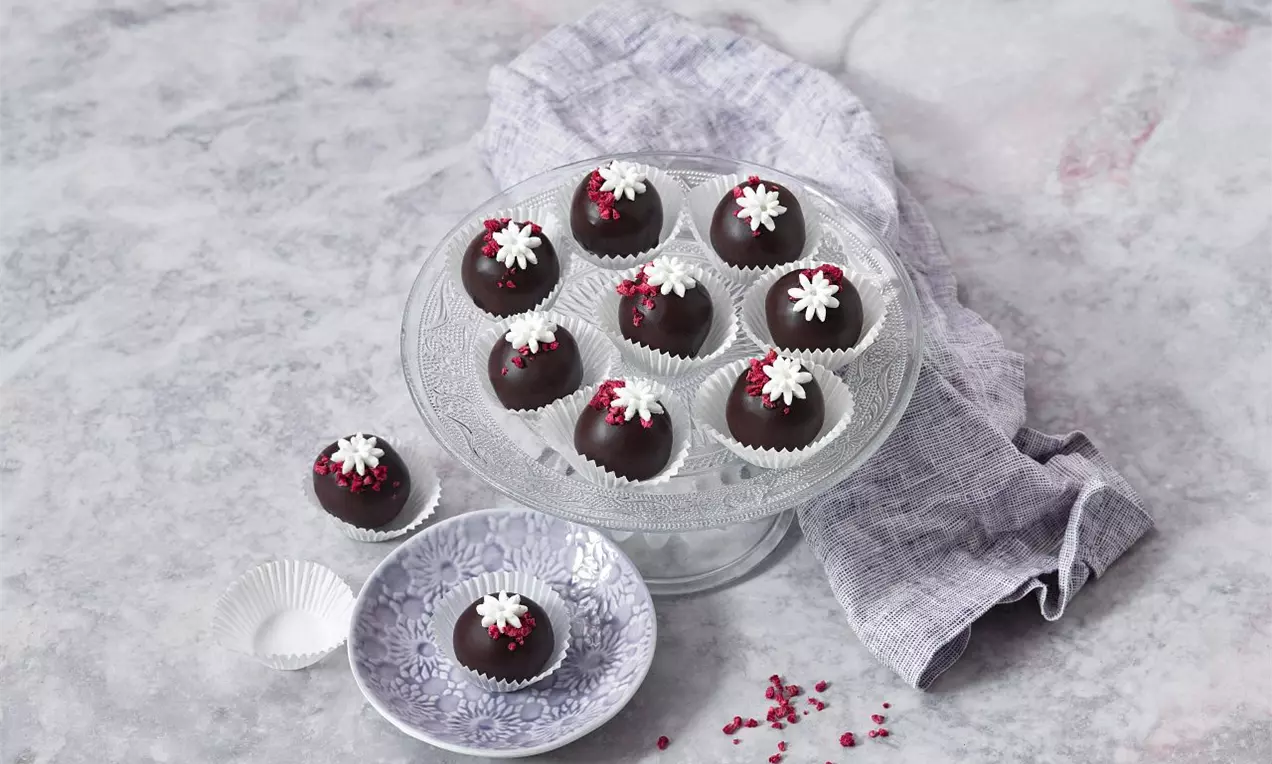 Čokoládové kulaté pralinky zdobené bílými květinami a drobnými červenými detaily, uspořádané na skleněném podnosu s látkovým ubrouskem