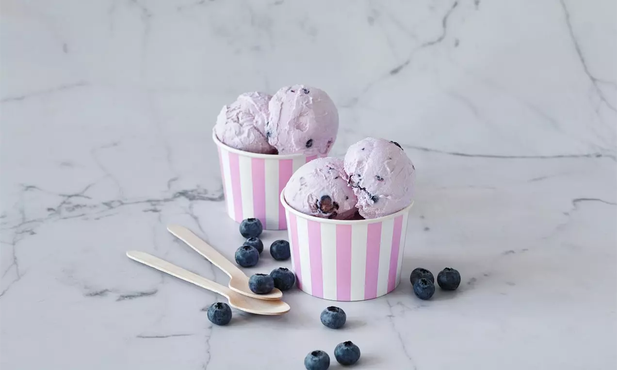 Světle fialová borůvková zmrzlina s jemnou krémovou texturou v pruhovaných kelímcích, obklopená čerstvými borůvkami na mramorovém podkladu